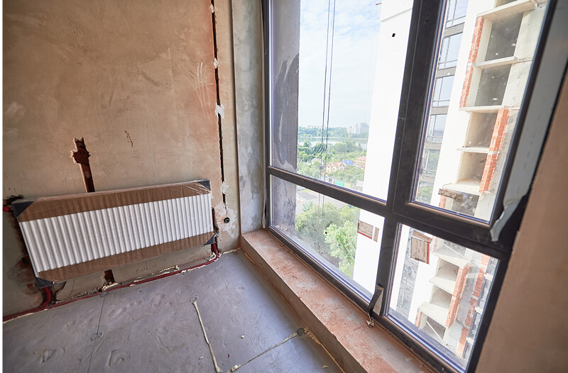 Unfinished room with exposed pipes, a covered radiator, and large windows overlooking a cityscape with buildings and greenery outside.
