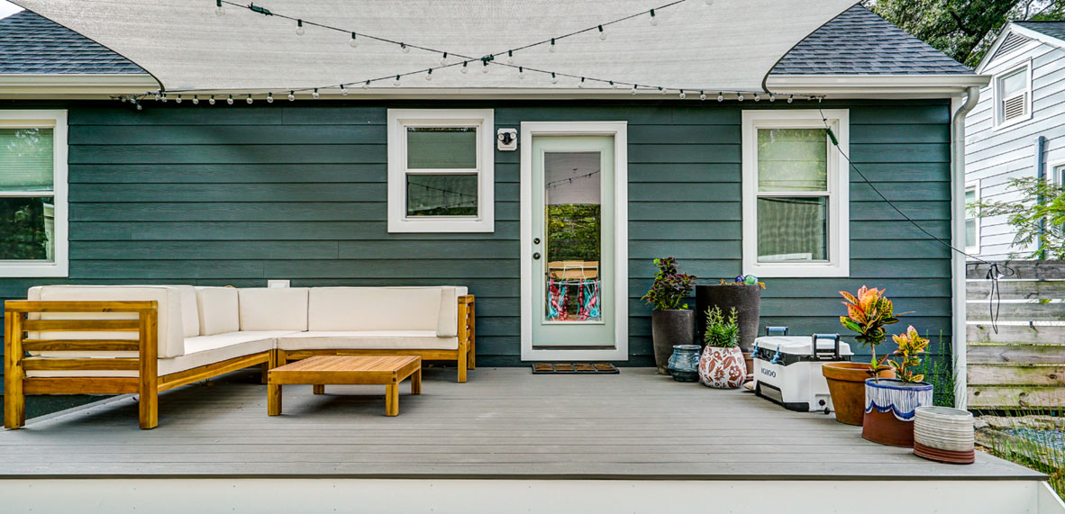 A backyard deck with a wooden outdoor sofa, coffee table, potted plants, a cooler, and string lights, attached to a green house with two windows and a door.