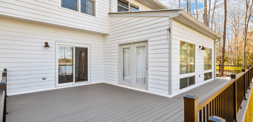 A spacious, modern outdoor deck attached to a white house with large windows, sliding glass doors, and a black railing, surrounded by trees.