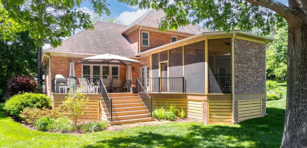 A brick house with a wooden deck, outdoor table and chairs, umbrella, and a screened porch, surrounded by green lawn and trees on a sunny day.