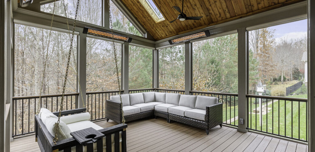 A screened-in porch with a vaulted wood ceiling, ceiling fan, heaters, a sectional sofa, a swing bench, and views of a green backyard.