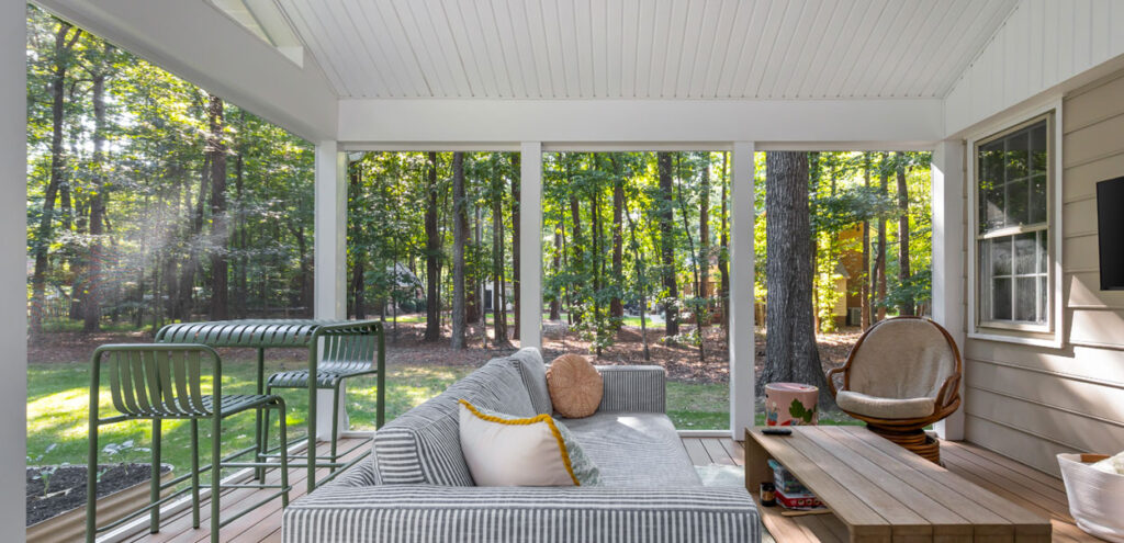 A screened-in porch with a striped sofa, a wooden coffee table, bar stools, and a chair, overlooking a wooded backyard.