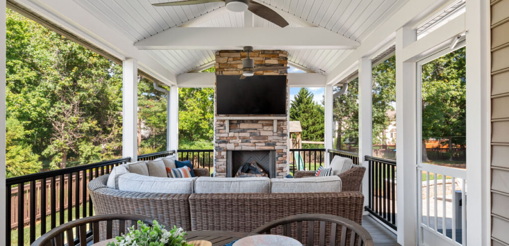 A covered patio with a wicker sectional sofa, coffee table, outdoor fireplace with a TV above, ceiling fans, and views of a green backyard.
