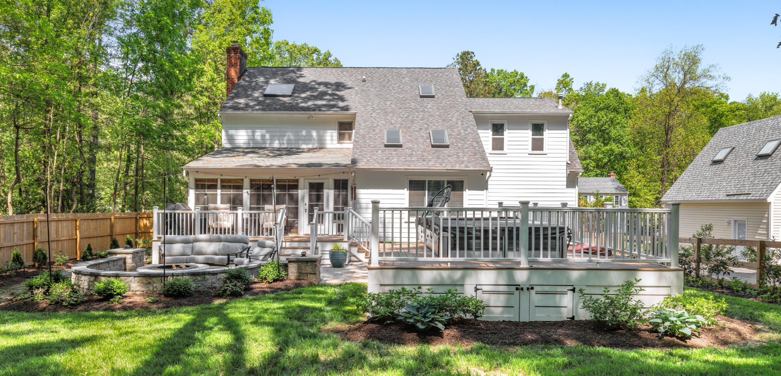 Back view of a two-story house with a deck, screened porch, and landscaped backyard featuring a fire pit area and surrounding trees.
