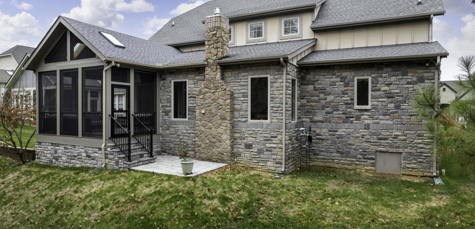 A stone exterior house with a screened porch, patio, chimney, several windows, and a sloped grassy yard.