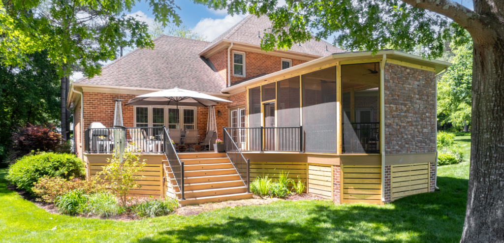 Brick house with a large wooden deck, outdoor seating area with an umbrella, and an attached screened porch, surrounded by green lawn and trees.