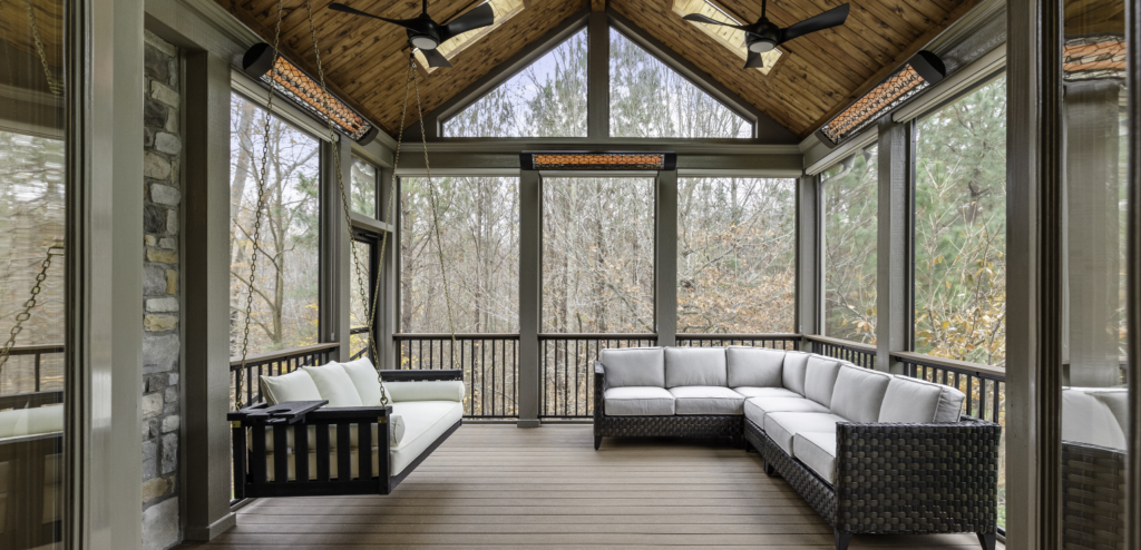 Screened-in porch with vaulted wooden ceiling, ceiling fans, a black porch swing with white cushions, and a wicker sectional sofa overlooking trees outside.