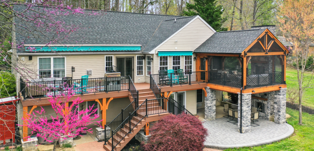 A two-story house with a large deck, outdoor seating, screened porch, and surrounding trees and landscaping.