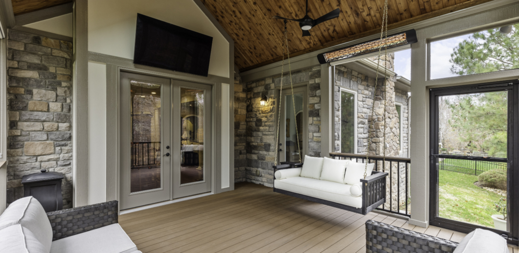 Covered patio with stone walls, glass doors, a ceiling fan, cushioned swing, wicker seating, and large windows overlooking a green backyard.