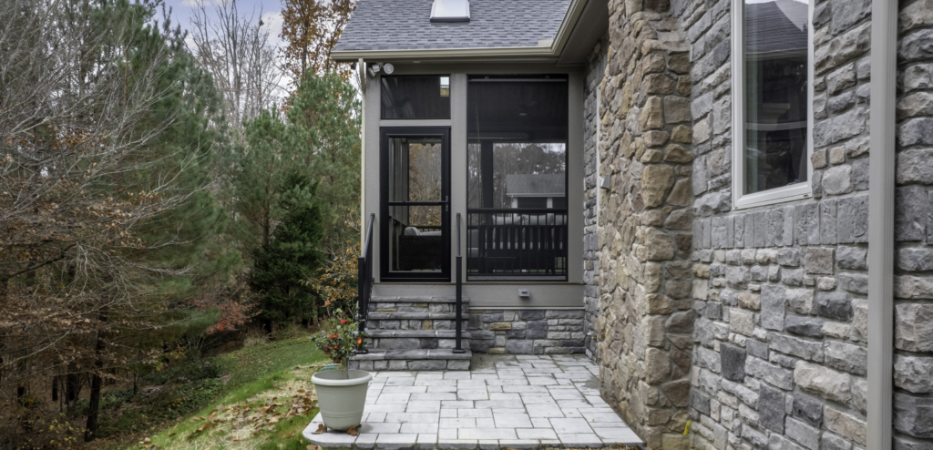 A stone patio with a potted plant leads to a screened porch attached to a stone house, surrounded by trees and greenery.