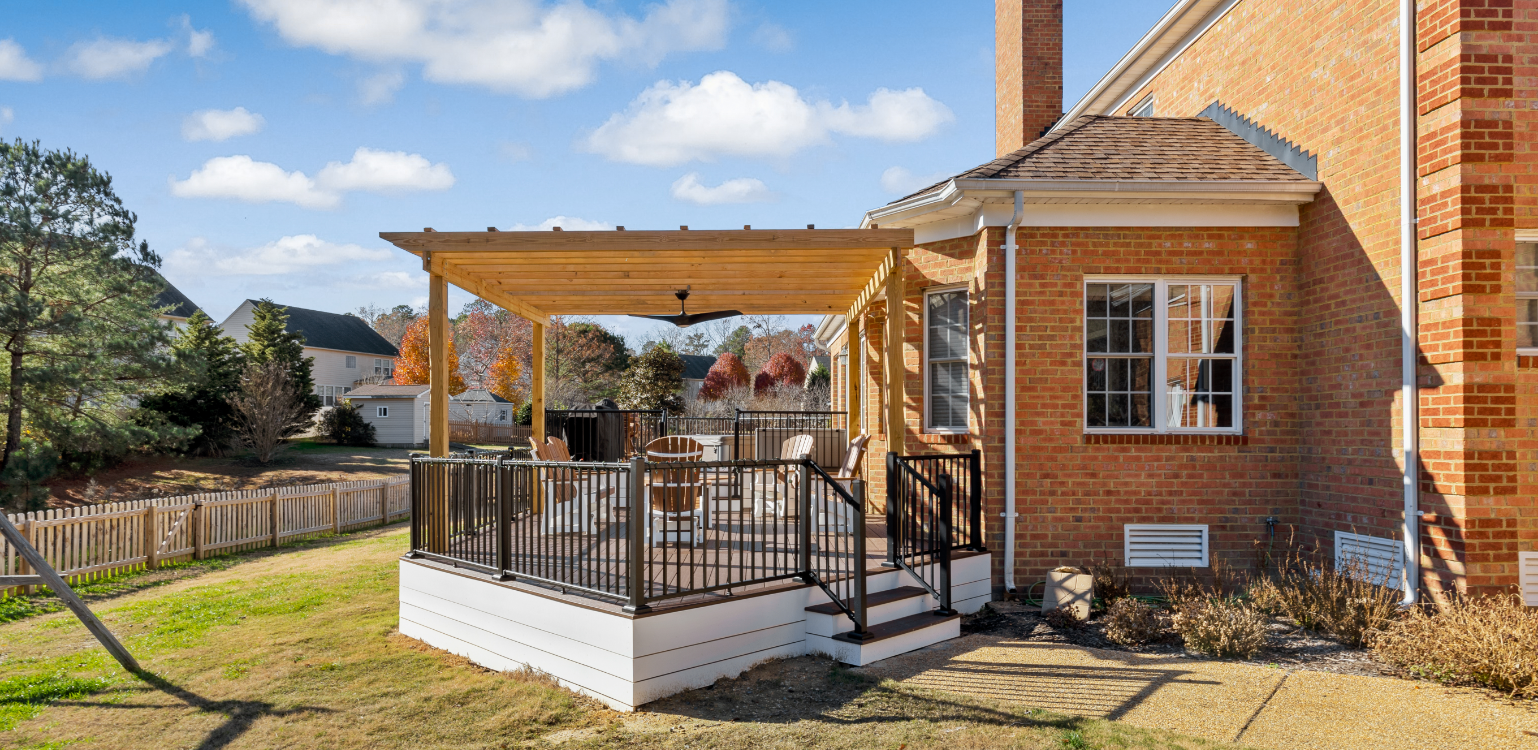 A brick house with a small covered deck featuring a black metal railing, outdoor furniture, and steps leading to a grassy yard.