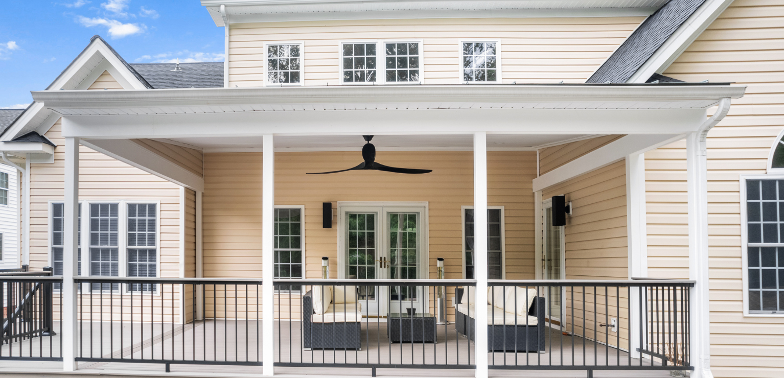A covered outdoor patio with a ceiling fan, black railing, and outdoor seating attached to a beige house with white trim and multiple windows.