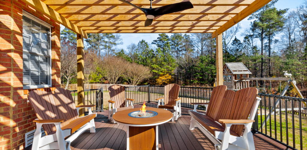 A covered patio with five wooden chairs around a fire pit table, ceiling fan above, and a view of a backyard with trees and a swing set.