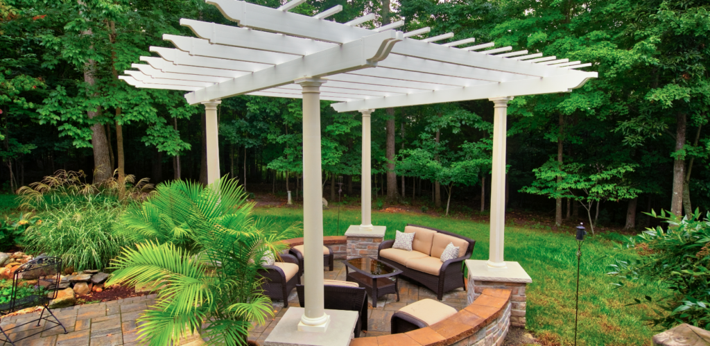 A patio seating area with cushioned chairs and a table is shaded by a white pergola, surrounded by greenery and trees.