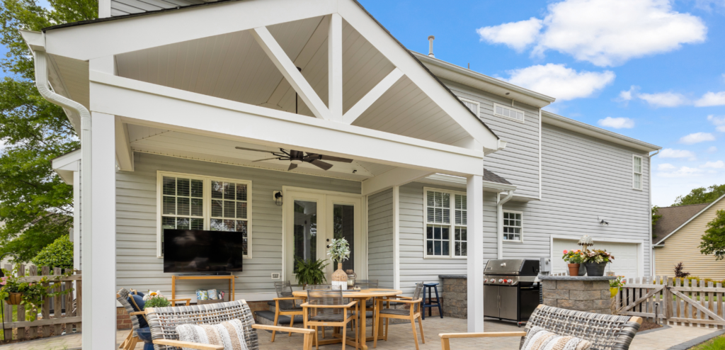Covered outdoor patio with seating area, TV, ceiling fan, dining table, grill, and potted plants, attached to a gray house on a sunny day.