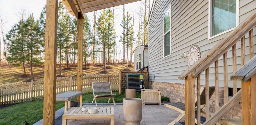 A backyard patio with wooden pergola, outdoor furniture, potted plants, and stairs leading to a beige house with a fenced yard and trees in the background.