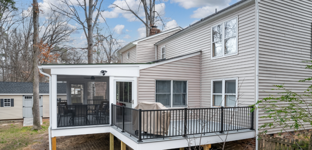 Back view of a house with beige siding, showing a screened porch and an open deck with black railings elevated above the yard.