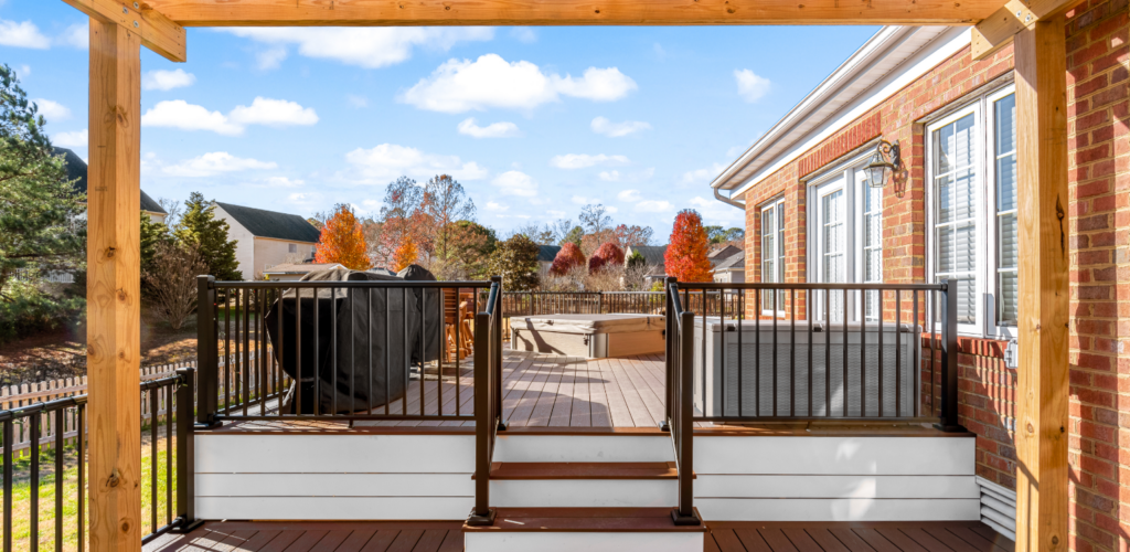 Wooden deck with black railings and a grill, attached to a brick house, overlooking a backyard with autumn trees.