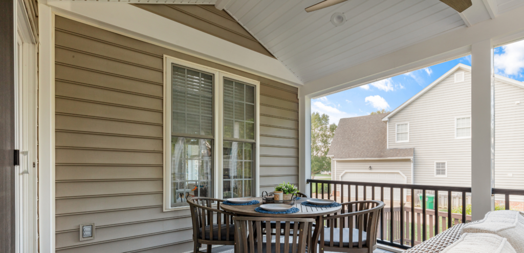 A covered porch with a round dining table set for four, wooden chairs, beige siding, and a view of a neighboring house.