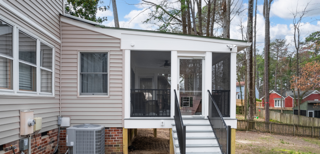 A screened-in porch with white stairs and black railing attached to a beige house, elevated above ground with a view of trees and neighboring homes.
