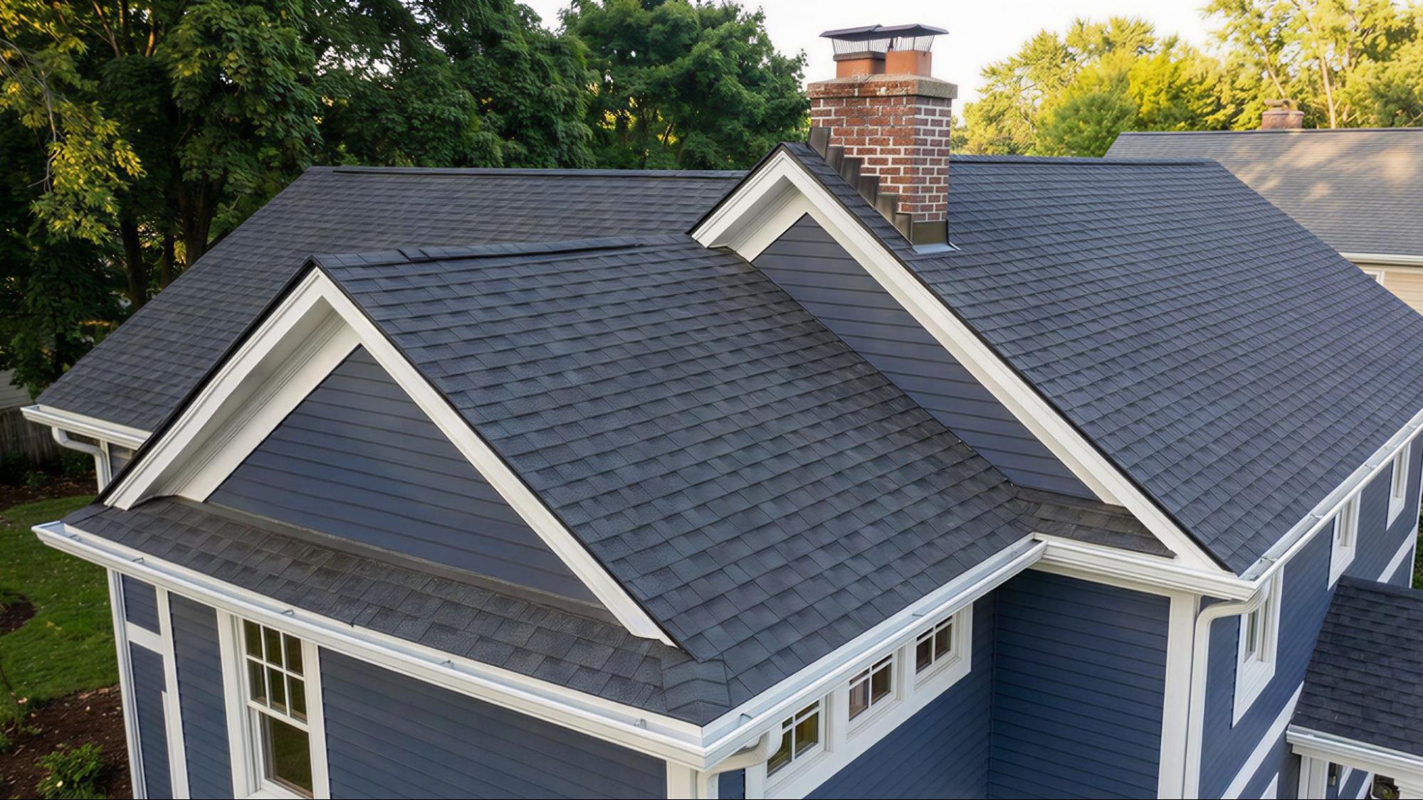 A dark gray shingle roof with a brick chimney on a blue house with white trim, surrounded by trees and sunlight.