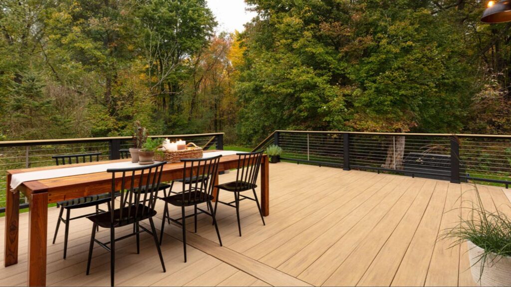 A wooden deck with a dining table and four chairs overlooks a forested area with trees showing early autumn foliage.