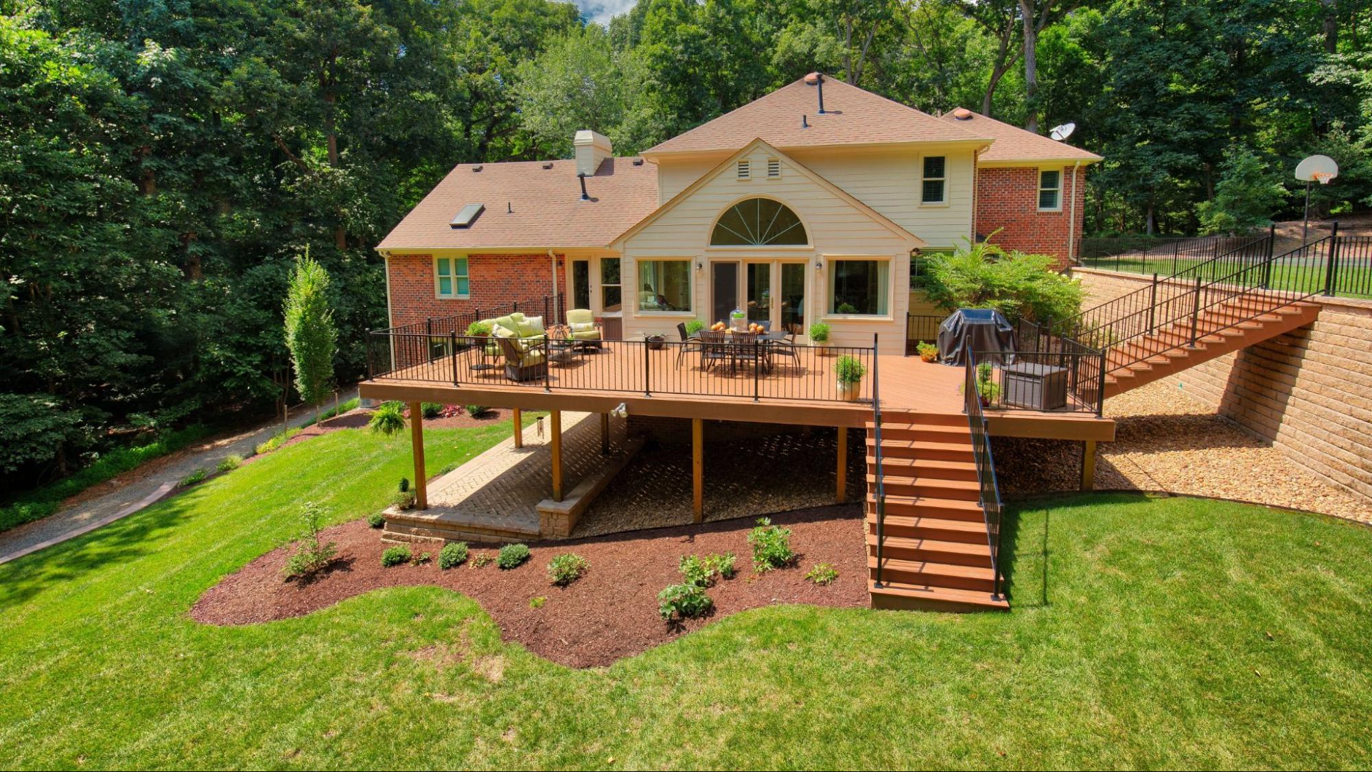 A two-story brick house with a spacious elevated deck, patio furniture, grill, landscaped yard, garden beds, and surrounding trees.