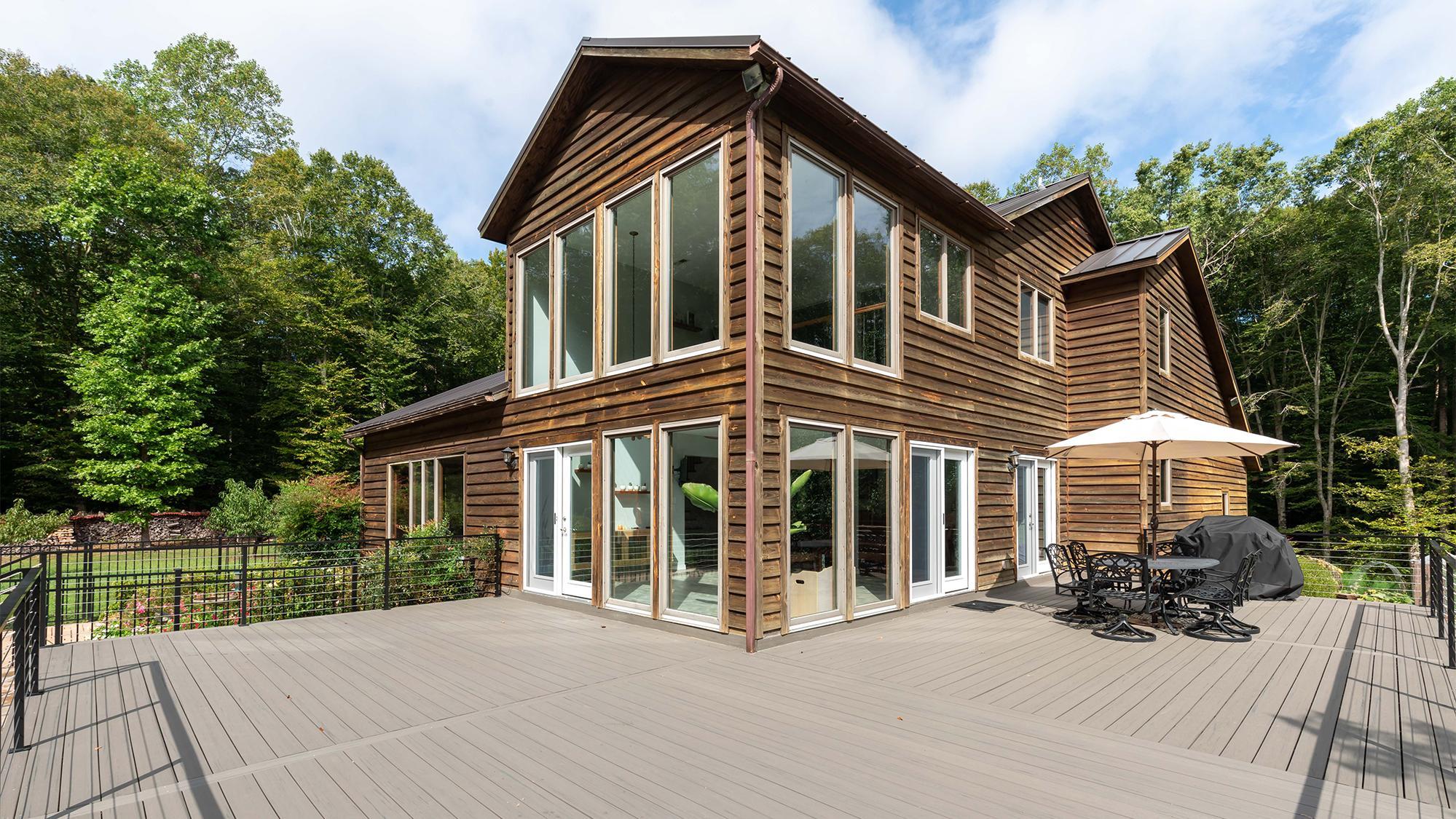 Modern two-story house with large windows and wood siding, surrounded by trees, featuring a spacious deck with outdoor seating and a grill.