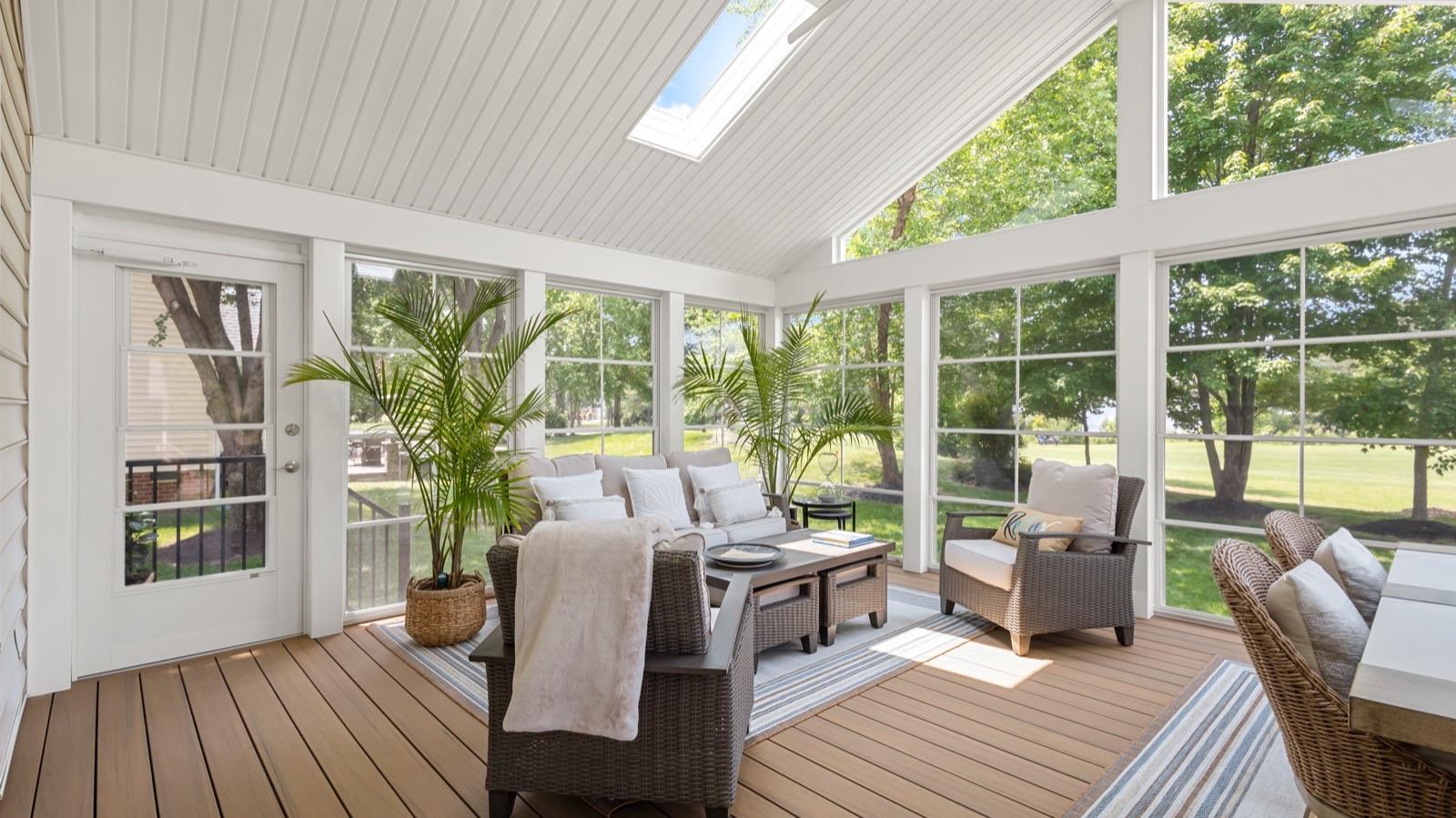 A bright, enclosed sunroom with large windows, wicker furniture, potted plants, and a wooden floor, overlooking a green lawn and trees outside.