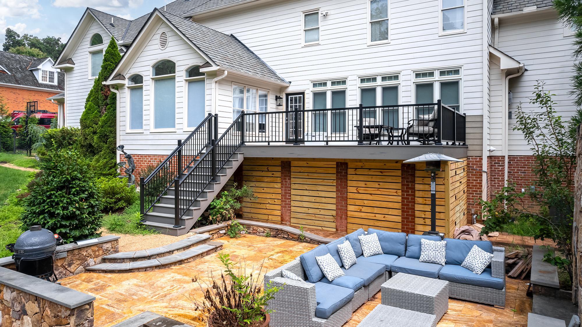 A modern backyard patio with a large outdoor sectional sofa, coffee tables, a grill, and a deck with stairs leading to the upper level of a white house.