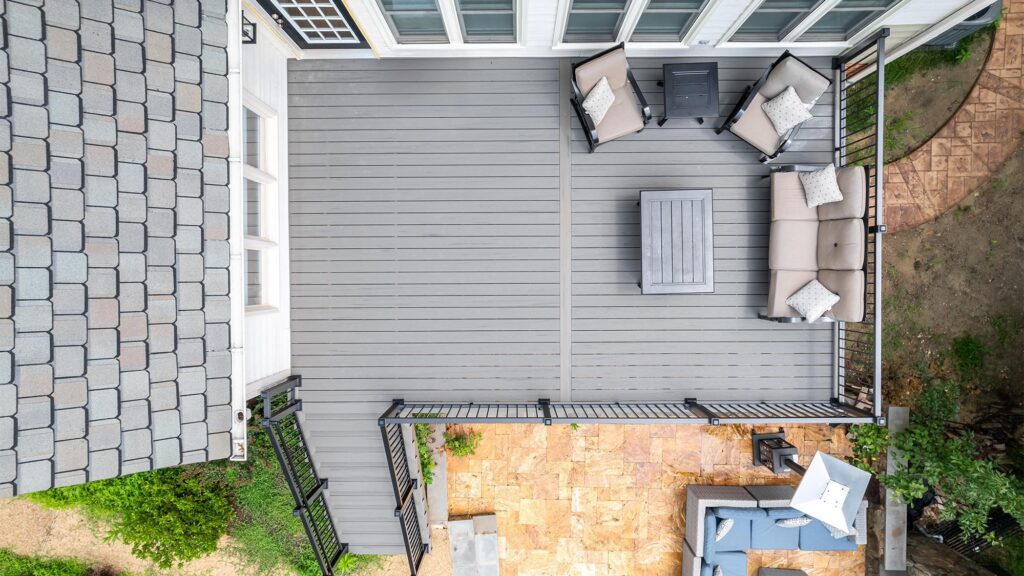 Aerial view of a modern backyard deck with gray flooring, outdoor seating, a coffee table, and railing, next to a stone patio area with more seating and an umbrella.
