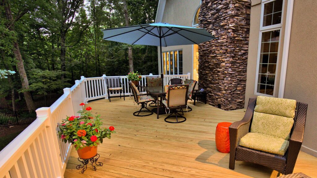 A wooden deck with patio furniture, including a table with chairs under a large umbrella, a wicker chair, potted plants, and a stone accent wall, overlooking a wooded area.