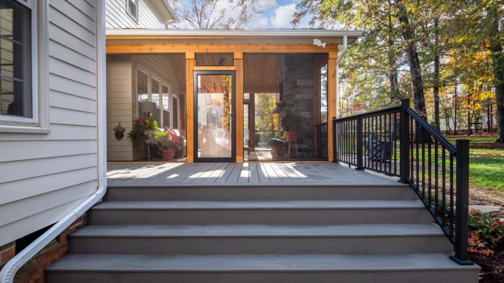 Wide wooden steps lead up to a screened-in porch with potted plants, attached to a light-colored house, surrounded by a grassy yard and trees.