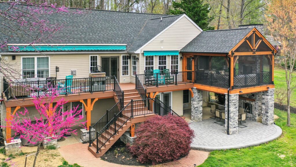 Two-story house with a large deck, screened porch, outdoor seating areas, and stone pillars, surrounded by trees and landscaped yard.