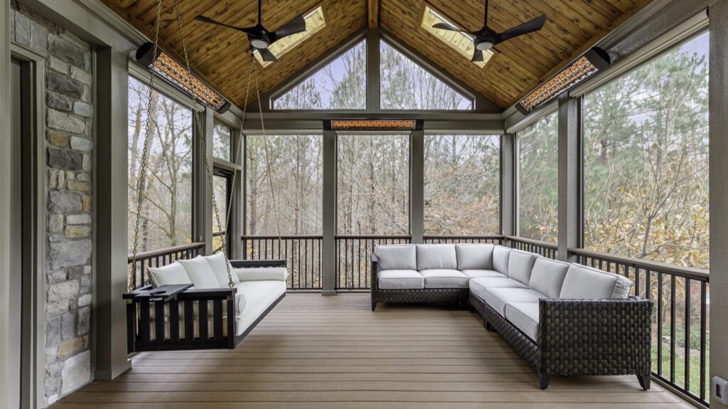 Screened-in porch with a wooden ceiling, ceiling fans, a white cushioned swing on the left, and an L-shaped wicker sofa on the right, overlooking trees.