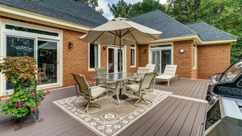 A brick house with a patio featuring a table, chairs, loungers, umbrella, patterned outdoor rug, grill, and potted flowers, surrounded by trees.