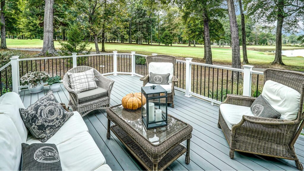Wicker patio furniture with cushions and decorative pillows arranged around a glass coffee table on a deck, overlooking a grassy lawn with trees.