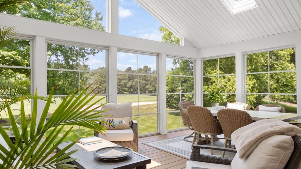A bright sunroom with large windows, wicker chairs around a table, and a potted plant, overlooking a green outdoor landscape.