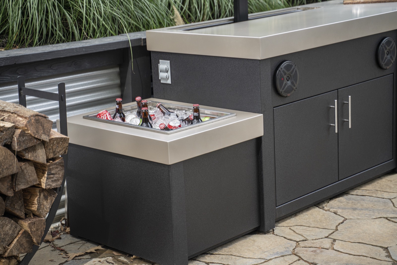 Built-in stainless ice trough filled with drinks in a dark outdoor kitchen — cabinetry sold by Deck Creations.
