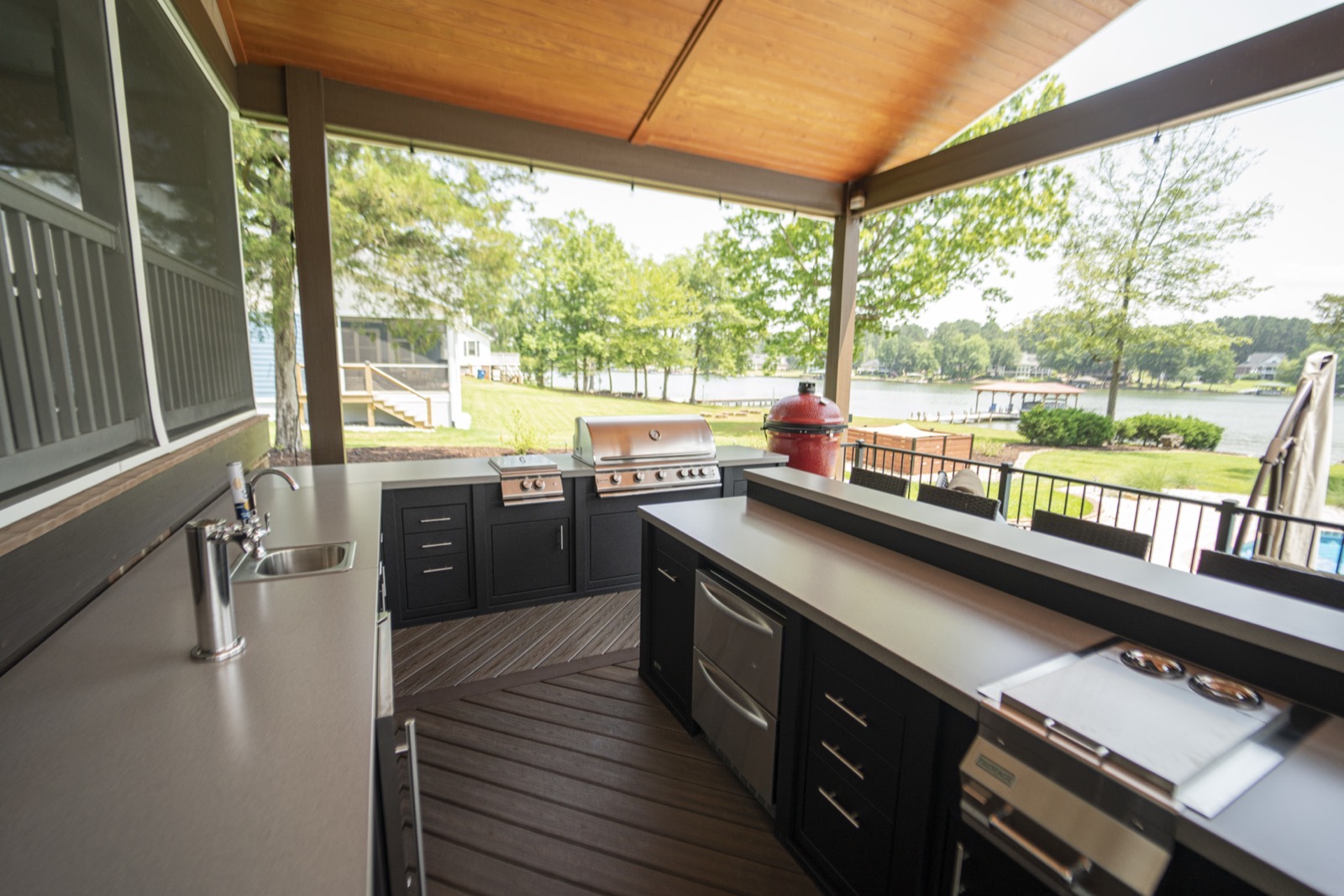 Dark outdoor kitchen with bar seating on a covered deck overlooking a lake — cabinetry available through Deck Creations.