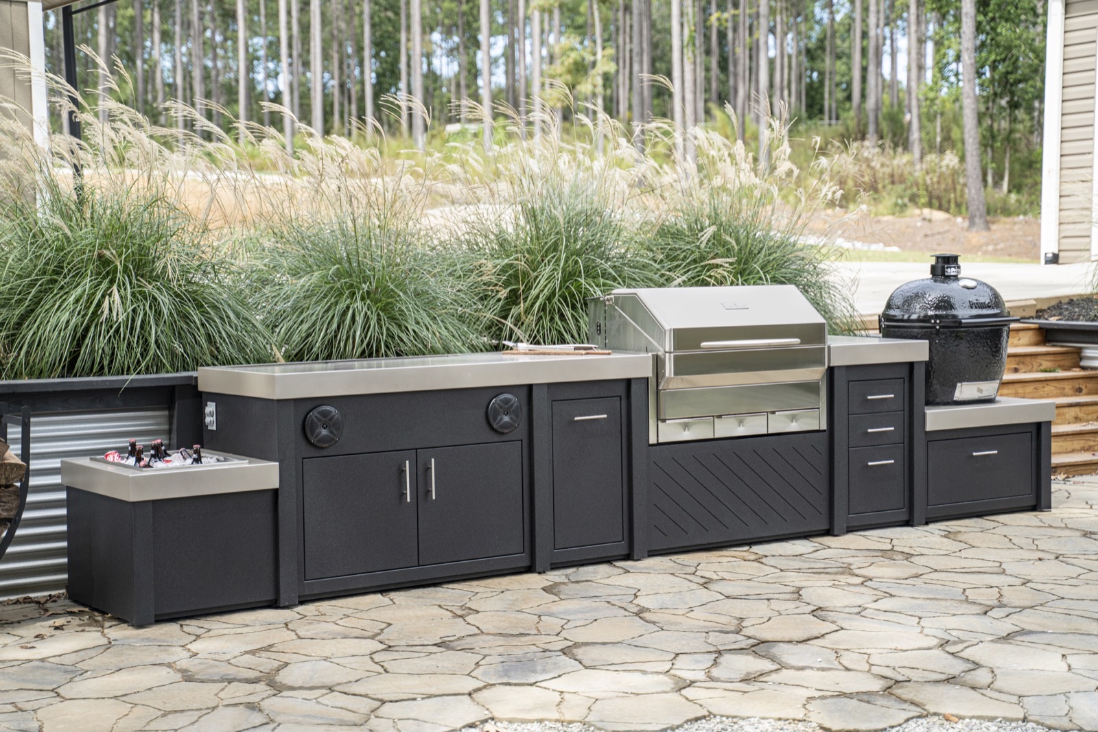 Dark entertainment kitchen with built-in speakers and a drink trough on a stone patio — from the cabinetry line Deck Creations offers.