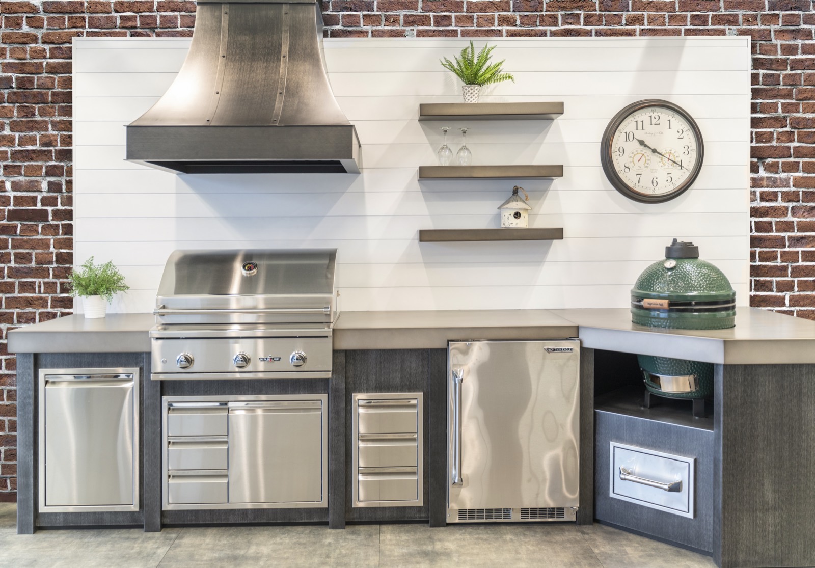 Outdoor kitchen with a metal vent hood, shiplap backsplash, open shelving and ceramic grill in gray cabinetry — sold by Deck Creations.