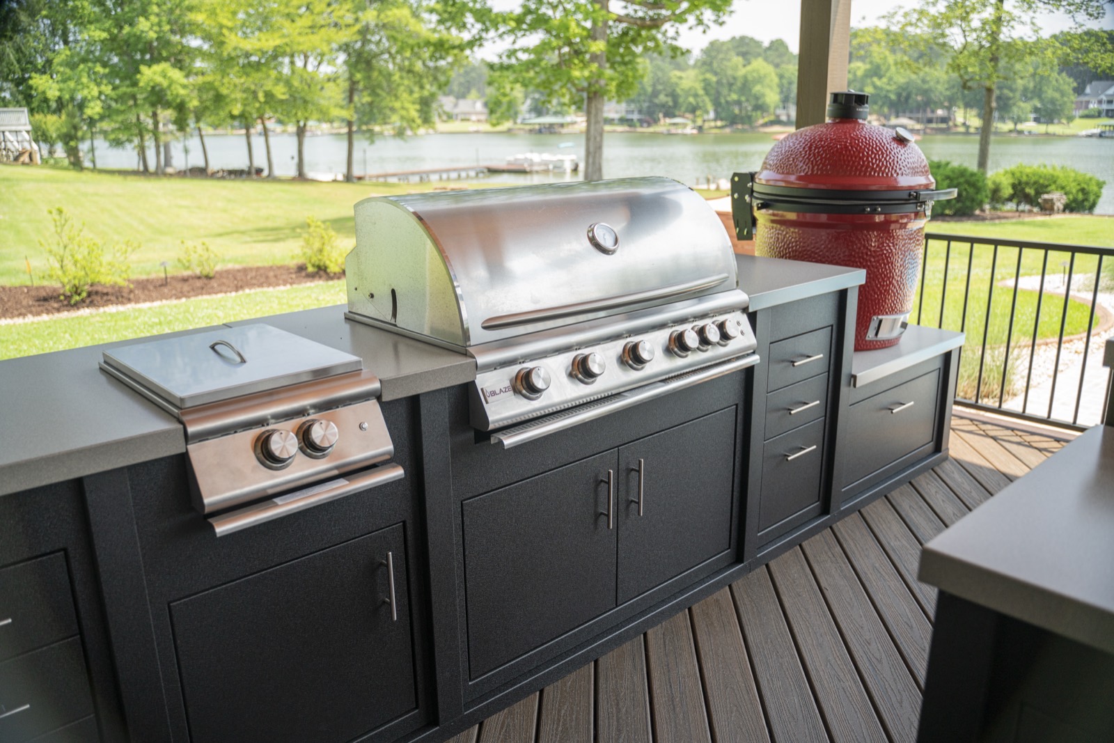 Dark outdoor kitchen with a stainless grill, side burner and red ceramic grill on a lakeside deck — cabinetry sold by Deck Creations.