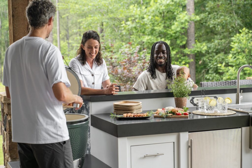Friends gathered around a white outdoor kitchen while the cook lifts the lid of a ceramic grill &mdash; custom cabinetry sold by Deck Creations.