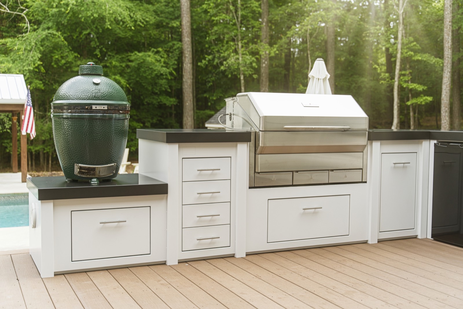 White outdoor kitchen with a green ceramic grill on a composite deck beside a pool — cabinetry available from Deck Creations.